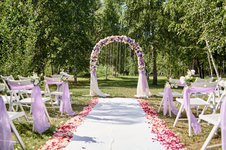 Place for wedding ceremony. Wedding arch decorated with cloth and flowers and chairs on each side of archway outdoors, copy space. Empty wooden chairs for guests on green grass. Wedding setup